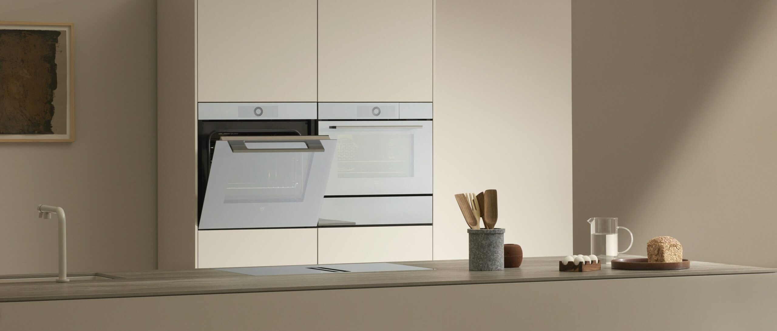 Moderne Küche mit Backofen, Arbeitsplatte, Spülbecken, Holzutensilien, Brot und Eiern in neutralen Tönen.