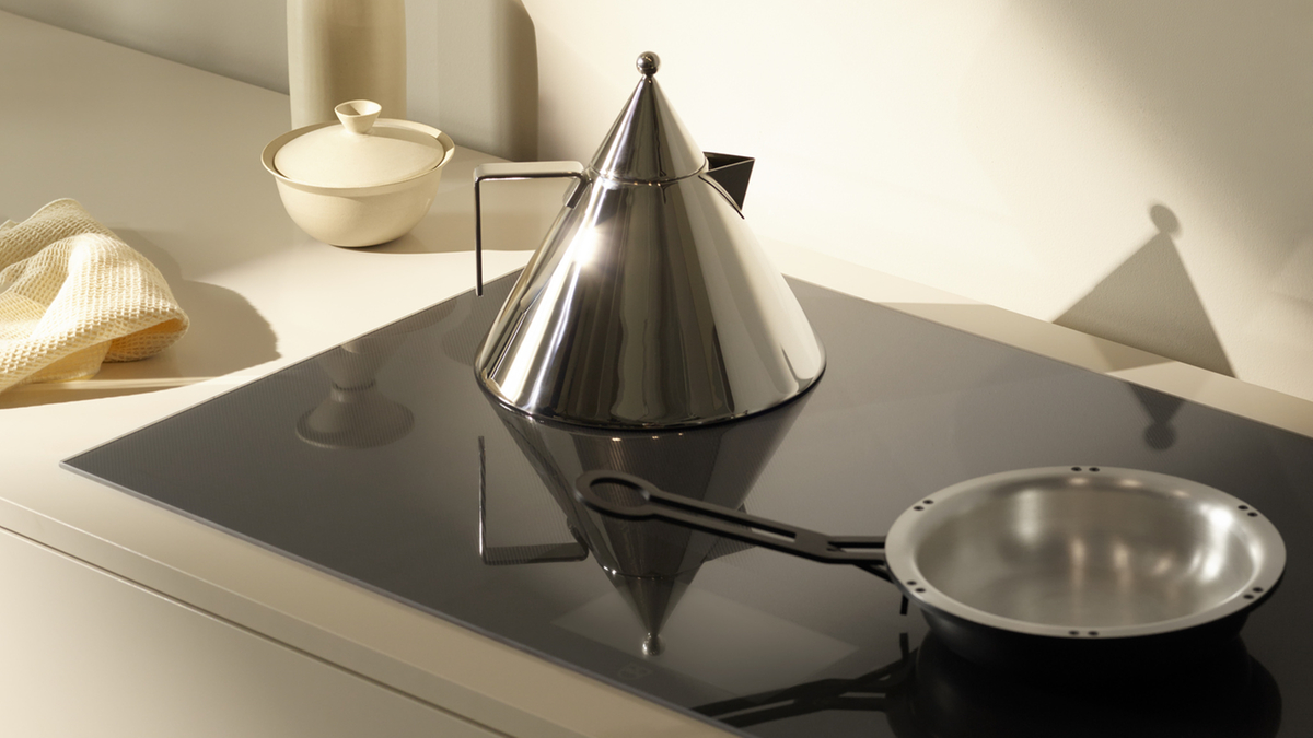 Minimalistische Küche mit glänzendem Wasserkocher, Pfanne auf Kochfeld und kleinem Topf im Hintergrund.