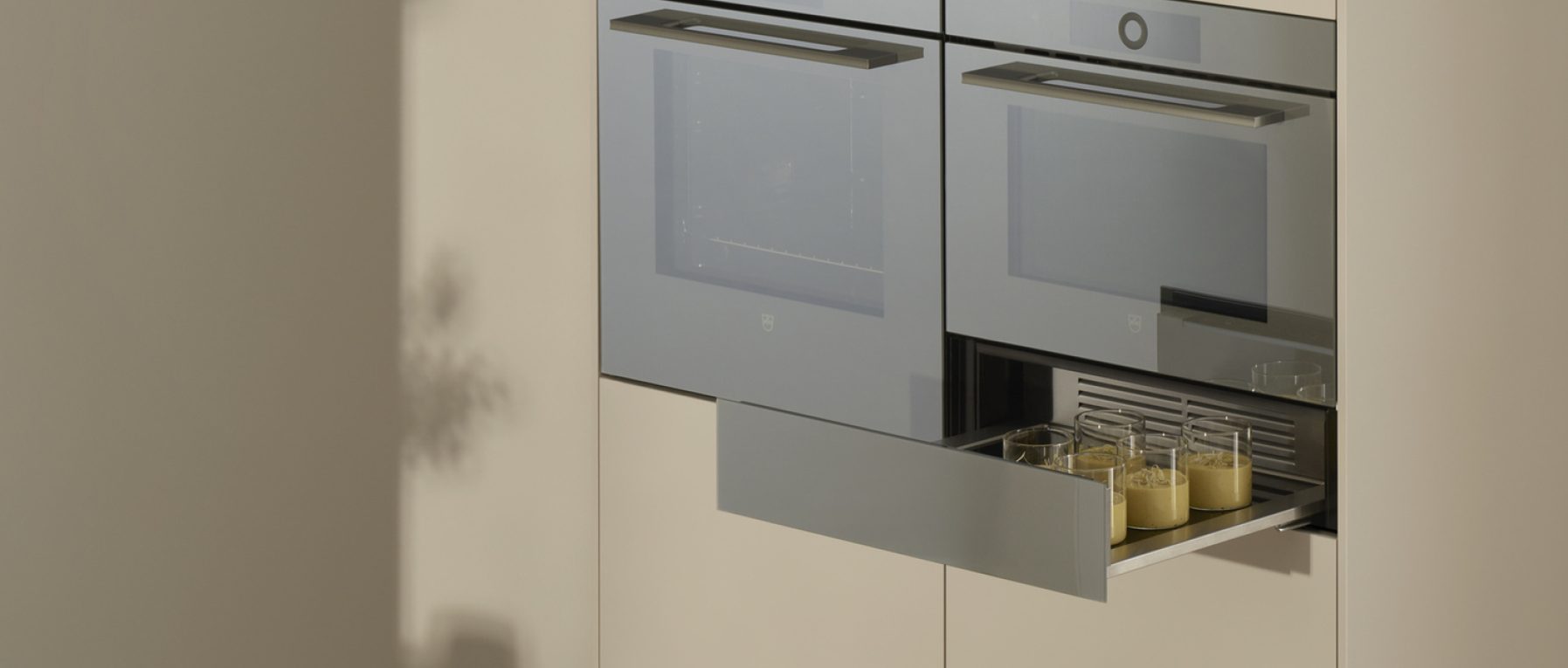 Moderne Küche mit zwei Einbaubacköfen, geöffnete Schublade mit Gläsern gelber Flüssigkeit darunter.